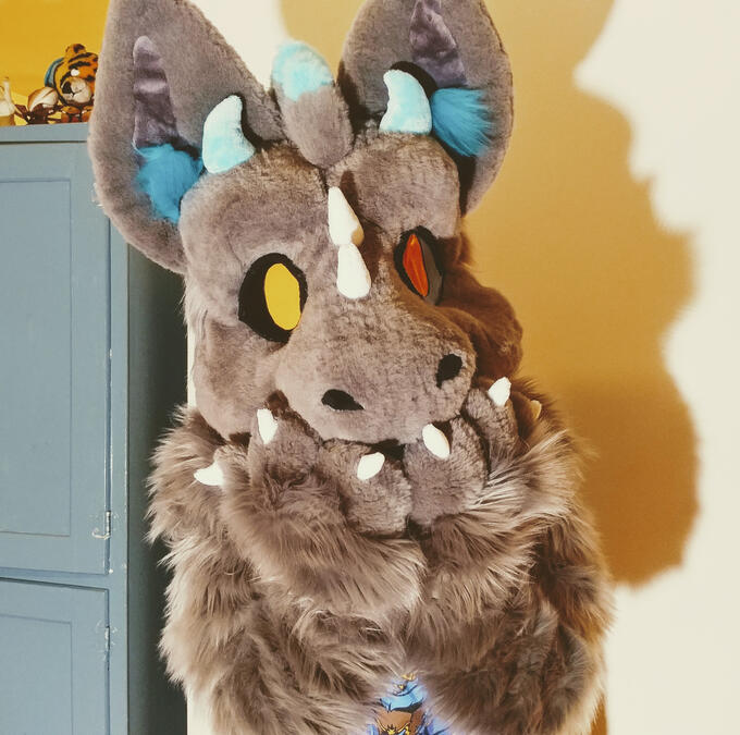 sky1 A person wearing the Sky fursuit, a grey dutch angel dragon. They are holding their paws over their mouth with their head slightly tilted.