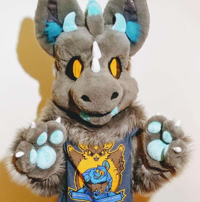 A person wearing the fursuit of Sky, a grey dutch angel dragon. They are standing with their paws raised slightly in front of them.