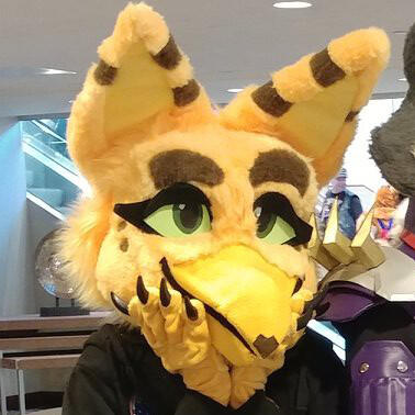 ani1 A person wearing the fursuit of Ani, a yellow avian creature. Their clawed hands are propped up beneath their chin.