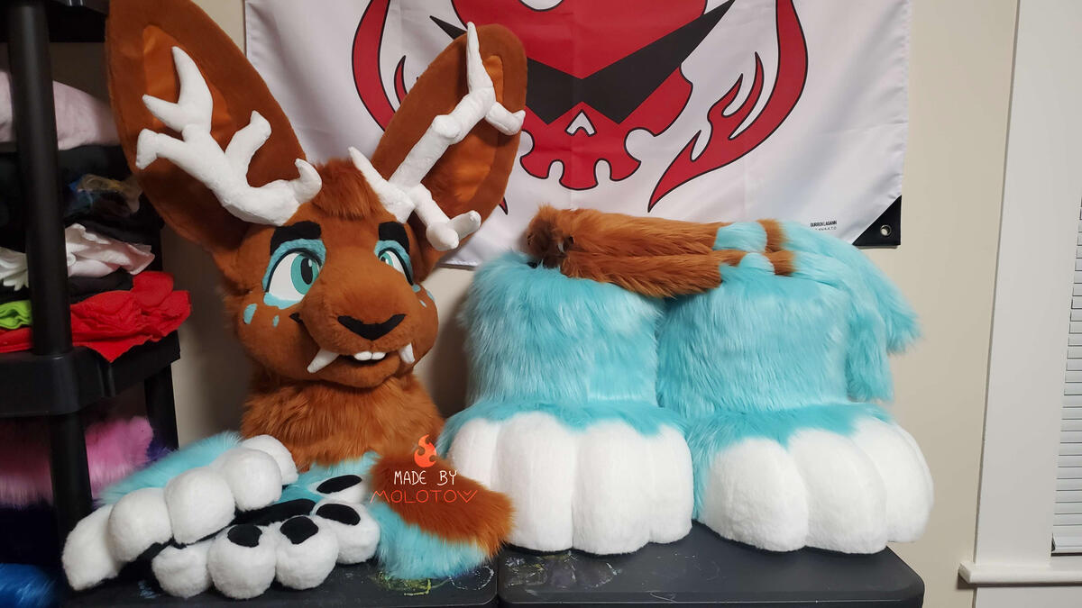 The fursuit head, handpaws, feetpaws, tail, and arm sleeves of Sienna, a wolpertinger character. He has a passive expression, two asymmetrical antlers, and saber teeth.